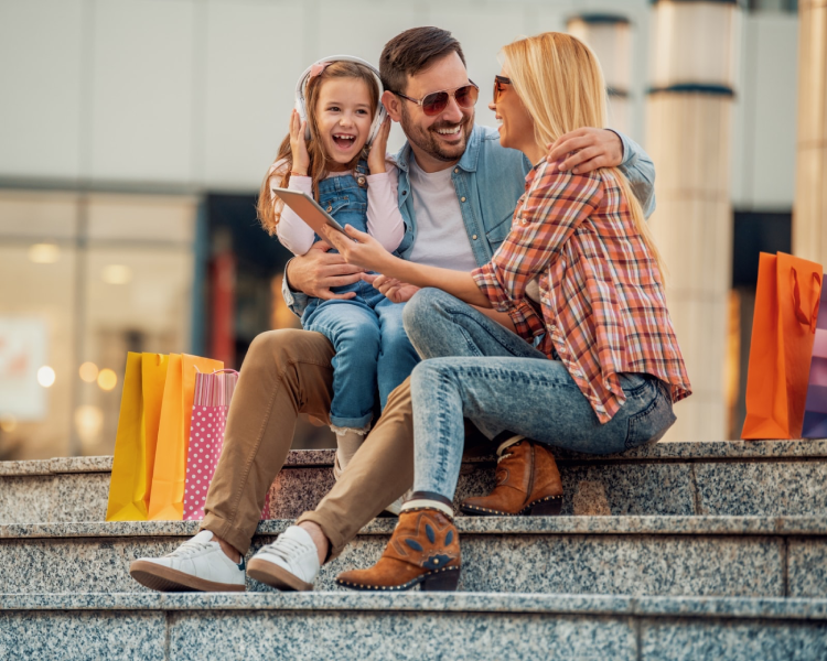 A mother, father, and child sitting and laughing on the steps of a shopping center with various shopping bags